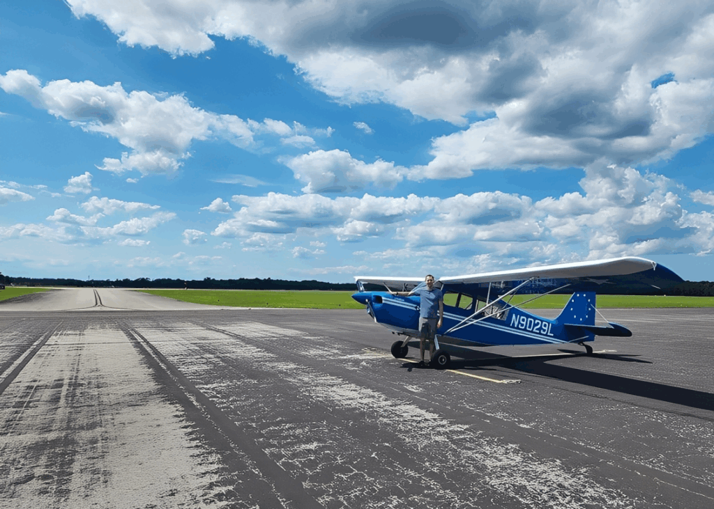 A pilot stands beside a blue and white aircraft on an airport tarmac under a partly cloudy sky